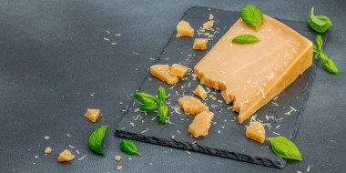 Modern tezgahta bir parça parmesan peyniri. Sarımsak, taze fesleğen, sağlıklı yemek için geleneksel İtalyan malzemeleri. Siyah taş zemin, düz zemin, pankart biçimi