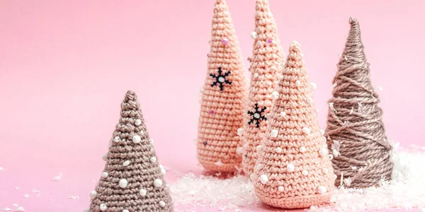 Handmade winter composition. Set of crocheted Christmas trees, artificial snow on pink background. Creative hobby concept, banner format