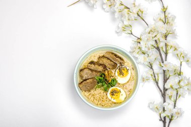 Japon eriştesi çorbalı Ramen. Sıcak yemek, ördek, yumurta, yeşillik. Beyaz arka plan, kiraz çiçekleri, düz yatak, üst manzara