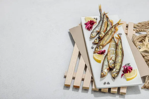 Smoked smelt with fresh lemon and herbs. Salted fish with marine decor. Trendy pallet, sea rope. Hard light, dark shadow, light stone concrete background, copy space