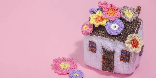 Handmade home spring decor concept. Creative crocheting, house figurine, traditional flowers. Festive greeting card, gentle pastel pink background, banner format
