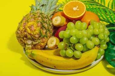 Bir dizi taze tropikal meyve. Olgun mango, ananas, portakal, üzüm ve muz. Sert ışık, koyu gölge, sarı arkaplan, kapat