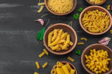 Makarna. Ahşap zemin üzerinde çeşitli pişmemiş makarna ve erişte çeşitleri. Klasik yemek hazırlamak için geleneksel İtalyan malzemesi, sebze ve otlar. Siyah tahtalar, düz yatış, üst manzara