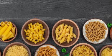 Makarna. Ahşap zemin üzerinde çeşitli pişmemiş makarna ve erişte çeşitleri. Klasik yemek hazırlamak için geleneksel İtalyan malzemesi, sebze ve otlar. Siyah panolar, düz döşeme, pankart biçimi