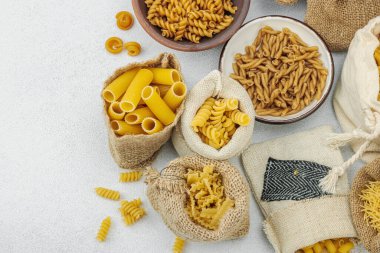 Makarna. Hafif taş zemin üzerinde çeşitli pişmemiş makarna ve erişte çeşitleri. Klasik yemek hazırlamak için geleneksel İtalyan malzemesi. El yapımı çuvallar, beyaz beton, üst görünüm