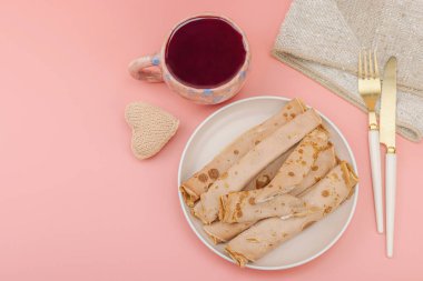 Bir fincan kokulu amber çayı ve kremalı ince krep. İyi ruh hali konsepti, rahat ev konforu. Pastel pembe arkaplan, üst görünüm