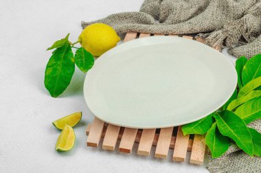 Yaratıcı mutfak konsepti. Boş tabak, olgun limon ve limon, taze yapraklar, modern stand ve eski peçete. Hafif taş arkaplan, düz yatış, kopyalama alanı
