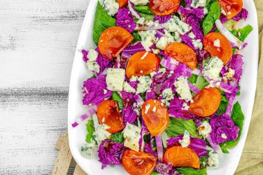 Healthy salad from purple Napa cabbage with persimmon, lettuce, Roquefort cheese and dressing. Fresh vegetables, delicious food. White wooden background, flat lay, top view clipart