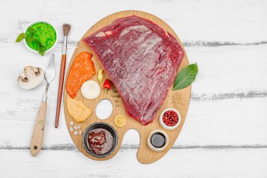 Çiğ biftek, çeşitli baharatlar, soslar ve ahşap ressamın paletinde yaratıcılıkla sıralanmış otlar içeren gurme yemek konsepti. Aşçılık ve aşçılık yaratıcılığı, beyaz ahşap arka plan, üst görünüm
