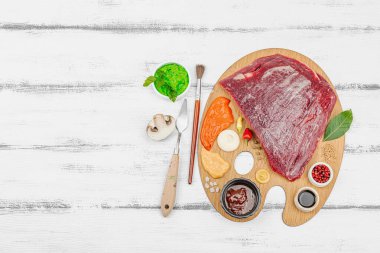 Çiğ biftek, çeşitli baharatlar, soslar ve ahşap ressamın paletinde yaratıcılıkla sıralanmış otlar içeren gurme yemek konsepti. Aşçılık ve aşçılık yaratıcılığı, beyaz ahşap arka plan, üst görünüm