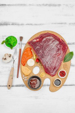 Çiğ biftek, çeşitli baharatlar, soslar ve ahşap ressamın paletinde yaratıcılıkla sıralanmış otlar içeren gurme yemek konsepti. Aşçılık ve aşçılık yaratıcılığı, beyaz ahşap arka plan, üst görünüm