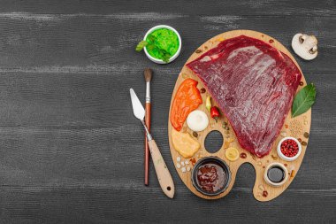 Çiğ biftek, çeşitli baharatlar, soslar ve ahşap ressamın paletinde yaratıcılıkla sıralanmış otlar içeren gurme yemek konsepti. Aşçılık ve aşçılık yaratıcılığı, siyah ahşap arka plan, üst görünüm