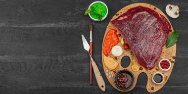 Çiğ biftek, çeşitli baharatlar, soslar ve ahşap bir ressamın paletinde yaratıcı bir şekilde sıralanmış otlarla dolu gurme yemek konsepti. Aşçılık ve aşçılık yaratıcılığı, siyah ahşap arka plan, afiş