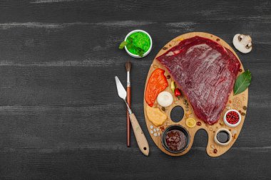 Çiğ biftek, çeşitli baharatlar, soslar ve ahşap ressamın paletinde yaratıcılıkla sıralanmış otlar içeren gurme yemek konsepti. Aşçılık ve aşçılık yaratıcılığı, siyah ahşap arka plan, üst görünüm