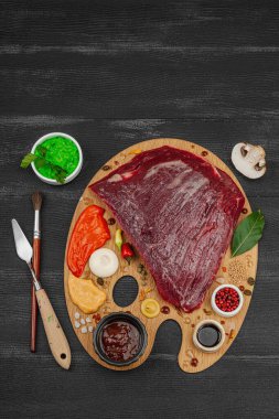 Çiğ biftek, çeşitli baharatlar, soslar ve ahşap ressamın paletinde yaratıcılıkla sıralanmış otlar içeren gurme yemek konsepti. Aşçılık ve aşçılık yaratıcılığı, siyah ahşap arka plan, üst görünüm