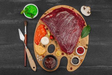 Çiğ biftek, çeşitli baharatlar, soslar ve ahşap ressamın paletinde yaratıcılıkla sıralanmış otlar içeren gurme yemek konsepti. Aşçılık ve aşçılık yaratıcılığı, siyah ahşap arka plan, üst görünüm