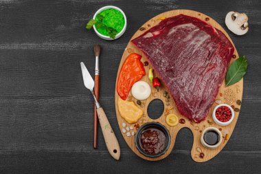 Çiğ biftek, çeşitli baharatlar, soslar ve ahşap ressamın paletinde yaratıcılıkla sıralanmış otlar içeren gurme yemek konsepti. Aşçılık ve aşçılık yaratıcılığı, siyah ahşap arka plan, üst görünüm