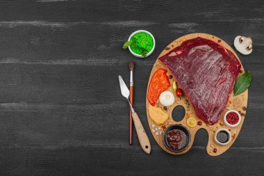 Çiğ biftek, çeşitli baharatlar, soslar ve ahşap ressamın paletinde yaratıcılıkla sıralanmış otlar içeren gurme yemek konsepti. Aşçılık ve aşçılık yaratıcılığı, siyah ahşap arka plan, üst görünüm
