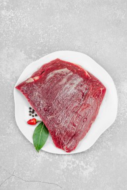 Beyaz seramik tabakta çeşitli baharatlarla taze çiğ biftek. Işık taşı arka planı. Et hazırlama, gurme pişirme ya da yemek tasarımı için mutfak konsepti.