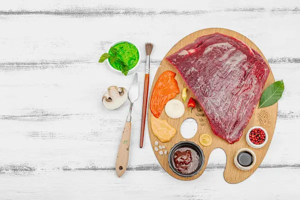Çiğ biftek, çeşitli baharatlar, soslar ve ahşap ressamın paletinde yaratıcılıkla sıralanmış otlar içeren gurme yemek konsepti. Aşçılık ve aşçılık yaratıcılığı, beyaz ahşap arka plan, üst görünüm