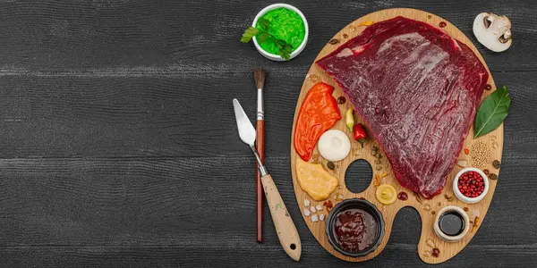 Çiğ biftek, çeşitli baharatlar, soslar ve ahşap bir ressamın paletinde yaratıcı bir şekilde sıralanmış otlarla dolu gurme yemek konsepti. Aşçılık ve aşçılık yaratıcılığı, siyah ahşap arka plan, afiş