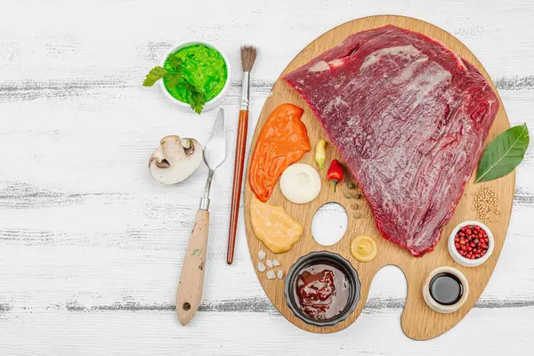 Çiğ biftek, çeşitli baharatlar, soslar ve ahşap ressamın paletinde yaratıcılıkla sıralanmış otlar içeren gurme yemek konsepti. Aşçılık ve aşçılık yaratıcılığı, beyaz ahşap arka plan, üst görünüm