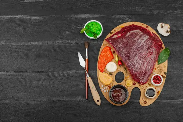 Çiğ biftek, çeşitli baharatlar, soslar ve ahşap ressamın paletinde yaratıcılıkla sıralanmış otlar içeren gurme yemek konsepti. Aşçılık ve aşçılık yaratıcılığı, siyah ahşap arka plan, üst görünüm