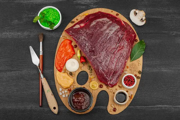 Çiğ biftek, çeşitli baharatlar, soslar ve ahşap ressamın paletinde yaratıcılıkla sıralanmış otlar içeren gurme yemek konsepti. Aşçılık ve aşçılık yaratıcılığı, siyah ahşap arka plan, üst görünüm