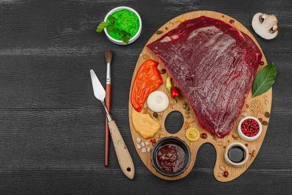 Çiğ biftek, çeşitli baharatlar, soslar ve ahşap ressamın paletinde yaratıcılıkla sıralanmış otlar içeren gurme yemek konsepti. Aşçılık ve aşçılık yaratıcılığı, siyah ahşap arka plan, üst görünüm