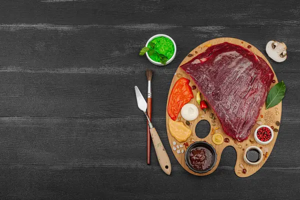 Çiğ biftek, çeşitli baharatlar, soslar ve ahşap ressamın paletinde yaratıcılıkla sıralanmış otlar içeren gurme yemek konsepti. Aşçılık ve aşçılık yaratıcılığı, siyah ahşap arka plan, üst görünüm