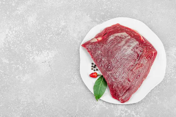 Beyaz seramik tabakta çeşitli baharatlarla taze çiğ biftek. Işık taşı arka planı. Et hazırlama, gurme pişirme ya da yemek tasarımı için mutfak konsepti.