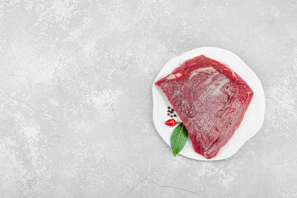 Beyaz seramik tabakta çeşitli baharatlarla taze çiğ biftek. Işık taşı arka planı. Et hazırlama, gurme pişirme ya da yemek tasarımı için mutfak konsepti.