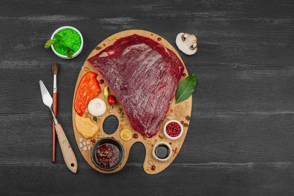 Çiğ biftek, çeşitli baharatlar, soslar ve ahşap ressamın paletinde yaratıcılıkla sıralanmış otlar içeren gurme yemek konsepti. Aşçılık ve aşçılık yaratıcılığı, siyah ahşap arka plan, üst görünüm