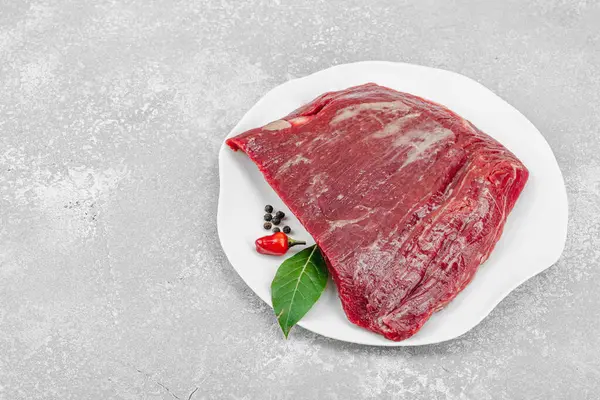 Beyaz seramik tabakta çeşitli baharatlarla taze çiğ biftek. Işık taşı arka planı. Et hazırlama, gurme pişirme veya yemek tasarımı için mutfak konsepti, kopyalama alanı