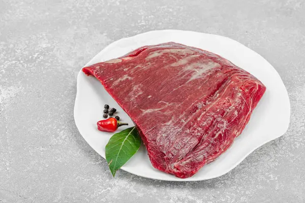 Beyaz seramik tabakta çeşitli baharatlarla taze çiğ biftek. Işık taşı arka planı. Et hazırlama, gurme pişirme ya da yemek tasarımı için mutfak konsepti.