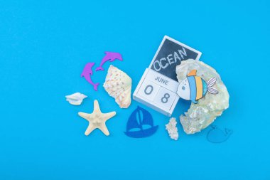 Dünya Okyanus Günü ve ekolojik farkındalık. Deniz kabukları, mercan, istiridye, yunus, tekne heykelciği ve 8 Haziran için takvim blokları içeren deniz temalı bir dekor. Eğitimsel resim, düz yatış, mavi arkaplan, üst görünüm