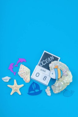 Dünya Okyanus Günü ve ekolojik farkındalık. Deniz kabukları, mercan, istiridye, yunus, tekne heykelciği ve 8 Haziran için takvim blokları içeren deniz temalı bir dekor. Eğitimsel resim, düz yatış, mavi arkaplan, üst görünüm