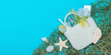 Dünya Okyanus Günü konsepti. Balık ağı, deniz yıldızı, deniz kabukları ve içi plastik, cam şişe, yosun gibi okyanus atıklarıyla dolu tekrar kullanılabilir keten çanta. Düz konum, mavi arkaplan, pankart biçimi