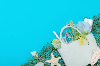Dünya Okyanus Günü konsepti. Balık ağı, deniz yıldızı, deniz kabukları ve içi plastik, cam şişe, yosun gibi okyanus atıklarıyla dolu tekrar kullanılabilir keten çanta. Düz konum, mavi arkaplan, üst görünüm