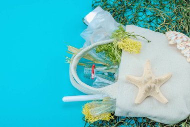 Dünya Okyanus Günü konsepti. Balık ağı, deniz yıldızı, deniz kabukları ve içi plastik, cam şişe, yosun gibi okyanus atıklarıyla dolu tekrar kullanılabilir keten çanta. Düz konum, mavi arkaplan, kapat