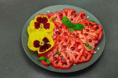 Aile yadigarı Coeur de Boeuf domatesi fesleğenli ve yenebilir çiçekli koyu tabakta. Temiz yemek, vejetaryen yaşam tarzı ve kırsal kesimde sunumlu tarladan sofraya yiyecek konsepti.