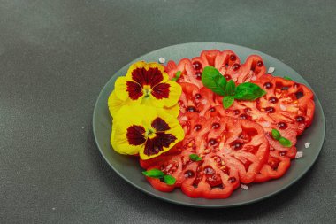 Aile yadigarı Coeur de Boeuf domatesi fesleğenli ve yenebilir çiçekli koyu tabakta. Temiz yemek, vejetaryen yaşam tarzı ve kırsal sunumlu tarladan sofraya yiyecek konsepti, kopyalama alanı