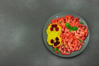 Aile yadigarı Coeur de Boeuf domatesi fesleğenli ve yenebilir çiçekli koyu tabakta. Temiz yemek, vejetaryen yaşam tarzı ve tarladan sofraya yiyecek konsepti.