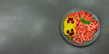 Aile yadigarı Coeur de Boeuf domatesi fesleğenli ve yenebilir çiçekli koyu tabakta. Temiz yemek, vejetaryen yaşam tarzı ve kırsal sunum, afiş formatlı çiftlikten sofraya yiyecek konsepti