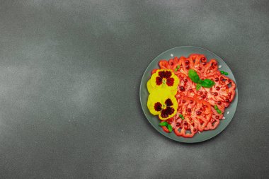 Aile yadigarı Coeur de Boeuf domatesi fesleğenli ve yenebilir çiçekli koyu tabakta. Temiz yemek, vejetaryen yaşam tarzı ve tarladan sofraya yiyecek konsepti.