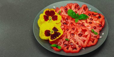 Aile yadigarı Coeur de Boeuf domatesi fesleğenli ve yenebilir çiçekli koyu tabakta. Temiz yemek, vejetaryen yaşam tarzı ve kırsal sunum, afiş formatlı çiftlikten sofraya yiyecek konsepti