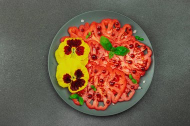 Aile yadigarı Coeur de Boeuf domatesi fesleğenli ve yenebilir çiçekli koyu tabakta. Temiz yemek, vejetaryen yaşam tarzı ve tarladan sofraya yiyecek konsepti.
