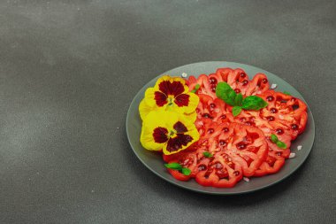 Aile yadigarı Coeur de Boeuf domatesi fesleğenli ve yenebilir çiçekli koyu tabakta. Temiz yemek, vejetaryen yaşam tarzı ve kırsal sunumlu tarladan sofraya yiyecek konsepti, kopyalama alanı