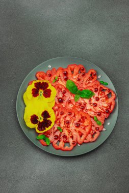 Aile yadigarı Coeur de Boeuf domatesi fesleğenli ve yenebilir çiçekli koyu tabakta. Temiz yemek, vejetaryen yaşam tarzı ve tarladan sofraya yiyecek konsepti.