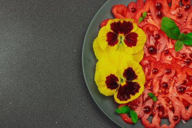 Aile yadigarı Coeur de Boeuf domatesi fesleğenli ve yenebilir çiçekli koyu tabakta. Temiz yemek, vejetaryen yaşam tarzı ve tarladan sofraya yiyecek konsepti.